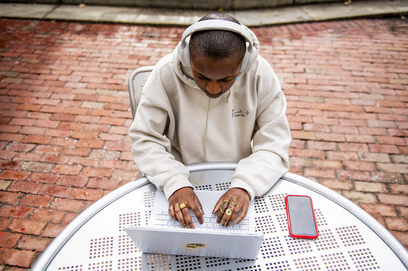 A black student works on a laptop outdoors, wearing a white hoodie and headphones.