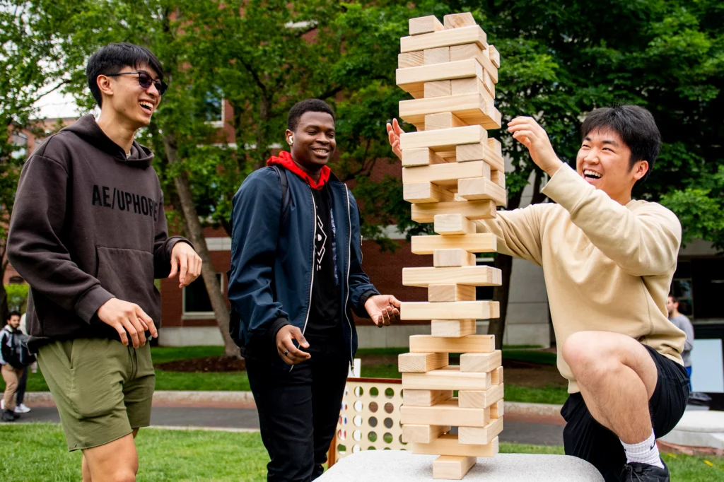 Three male students, two asian and one black, playing oversized Jenga outside.