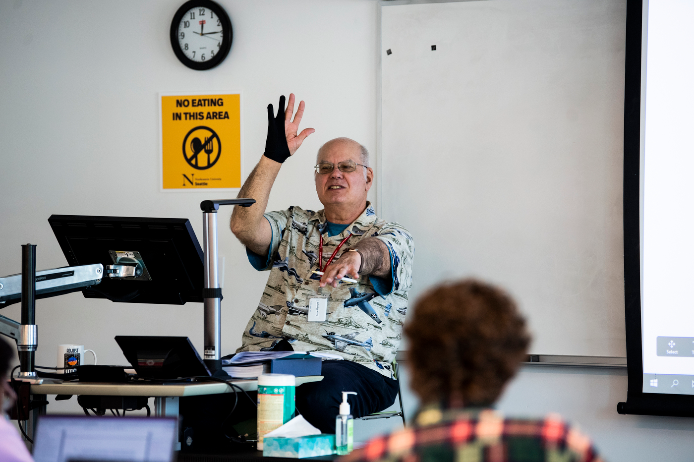 A white male faculty member raises his hand at the front of the class while holding a stylus pen forward in the other hand.